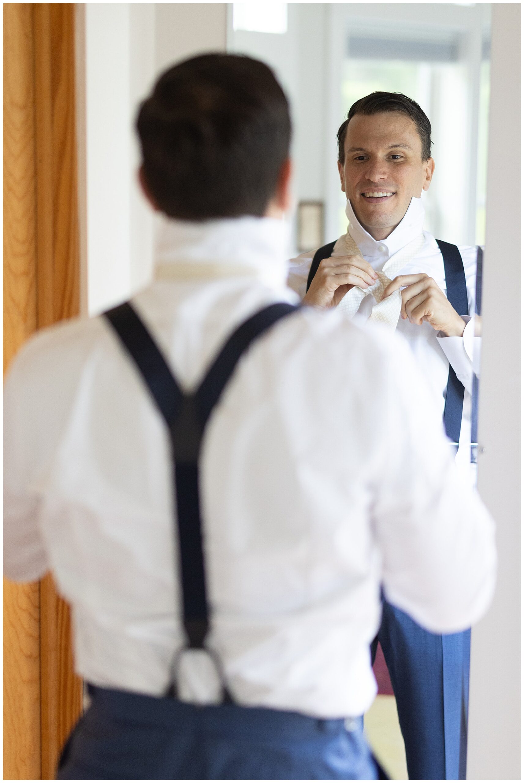 groom looks in the mirror as he ties his white tie on his wedding day suit 