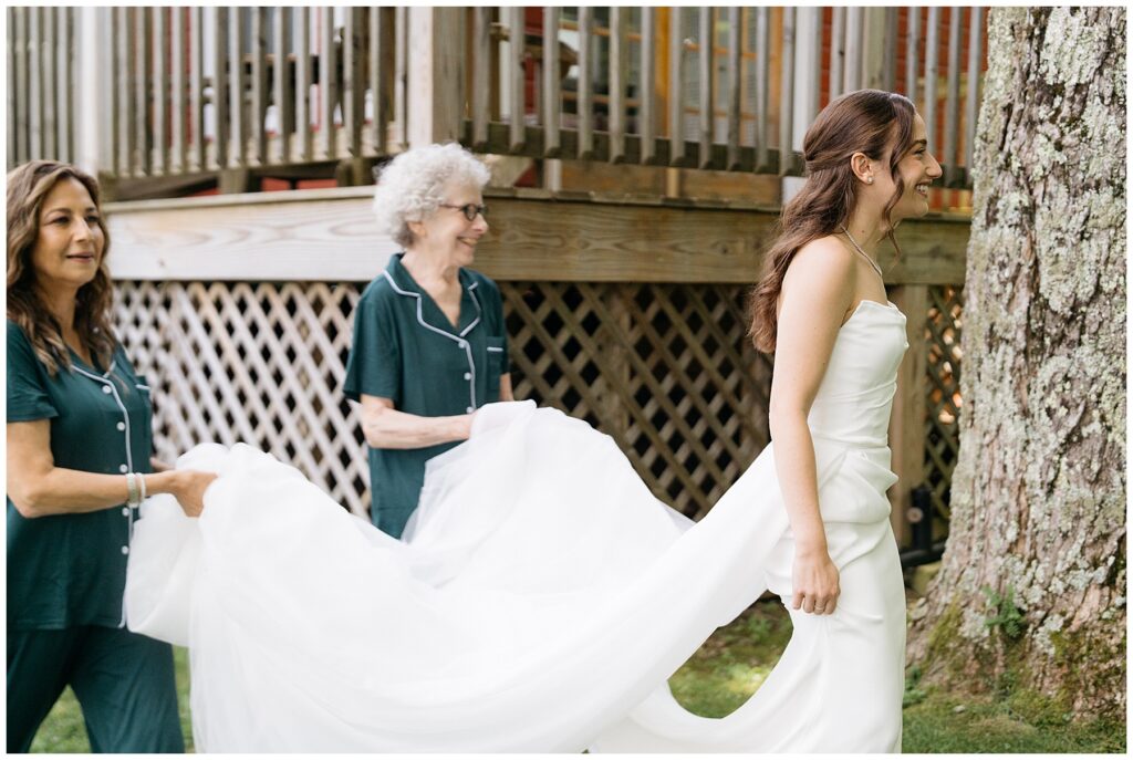 the bride smiles as she walks in her white wedding dress as her mother and grandmother walk behind her carrying the train wearing green pajamas 