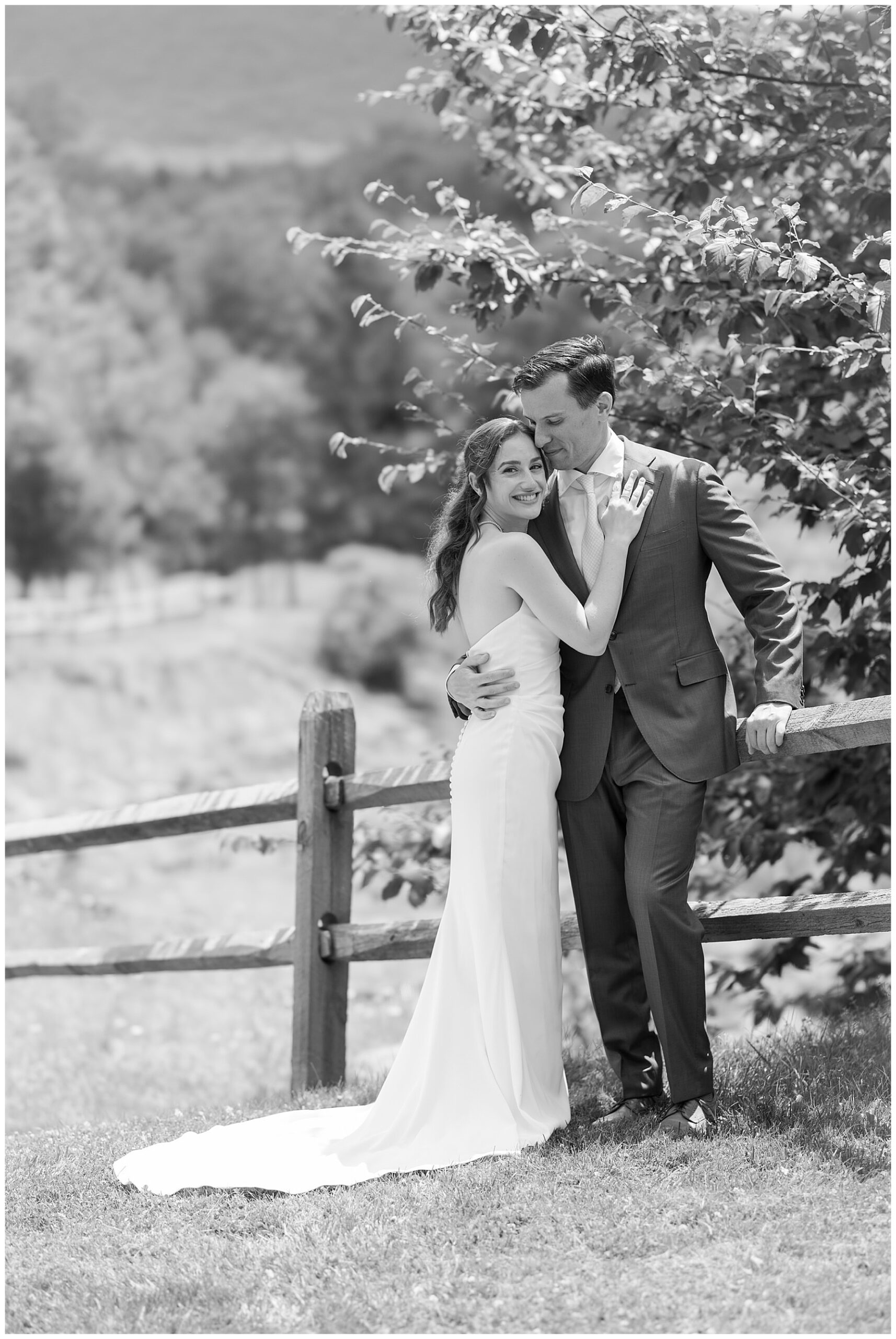 a black and white photo of the bride smiling at the camera as the groom holds her waist and leans into her