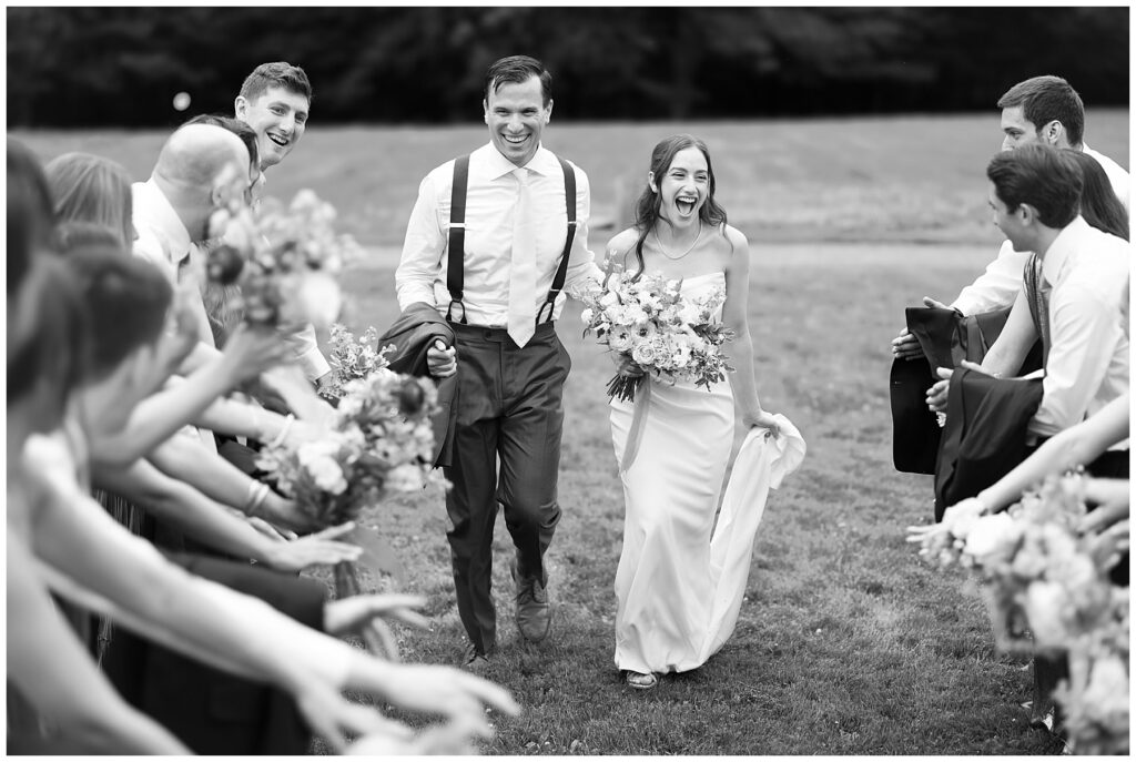 a black and white photo of the bride and groom walking down the row of their bridesmaids and groomsmen cheering them on before the wedding ceremony 