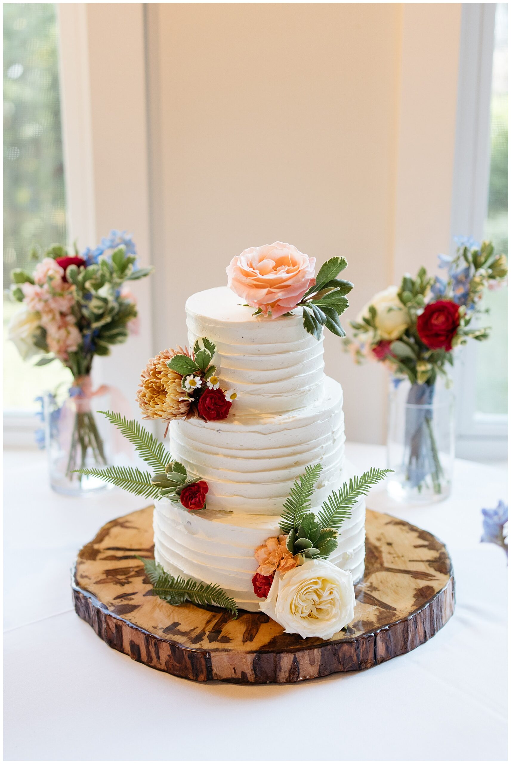 a white wedding cake covered in pink and orange flowers and green leaves sitting on top of a wood platter 