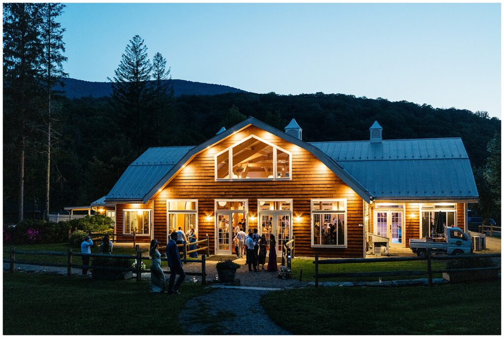 an exterior photo of the barn reception space at dusk with all the lights on and guests mingling outside of the full moon resort wedding